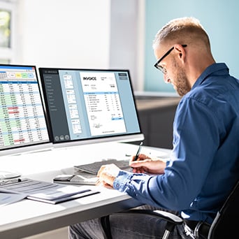 man sitting at a desk with two computer monitors working on paperwork 